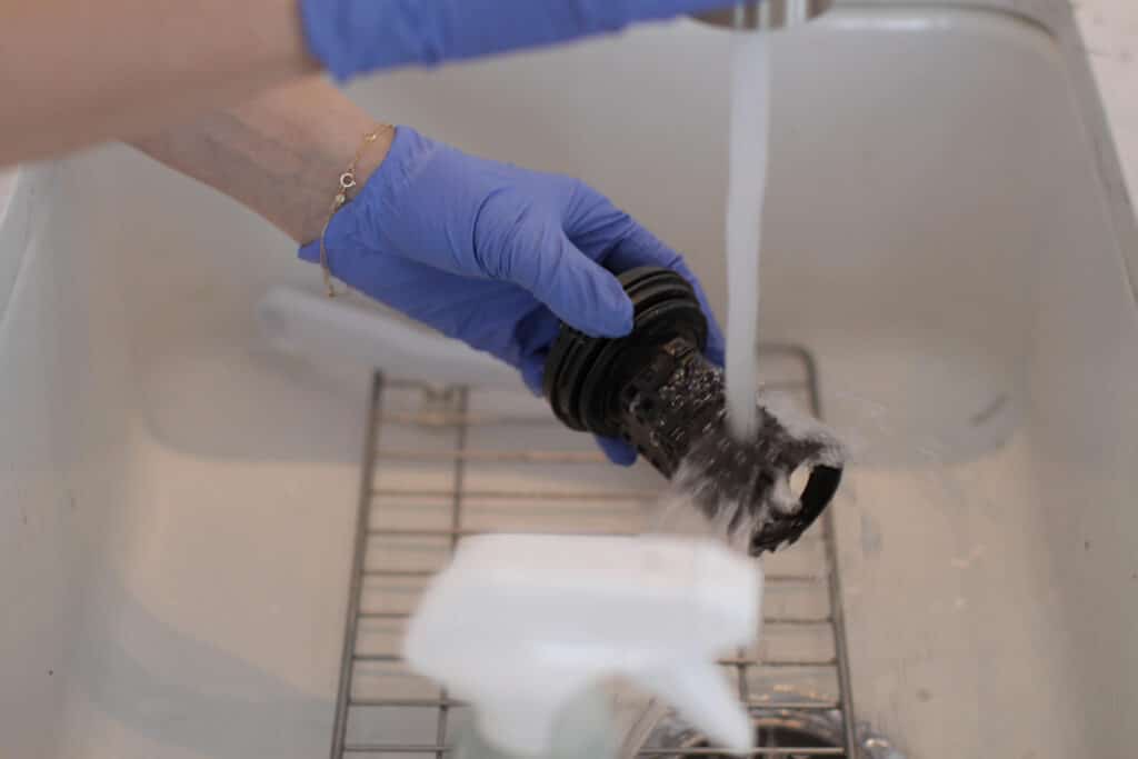 Photo 2025-02-04 11 31 29 AM - House Work A person wearing purple gloves rinses the dirty washing machine filter under running water in a sink to clean it.