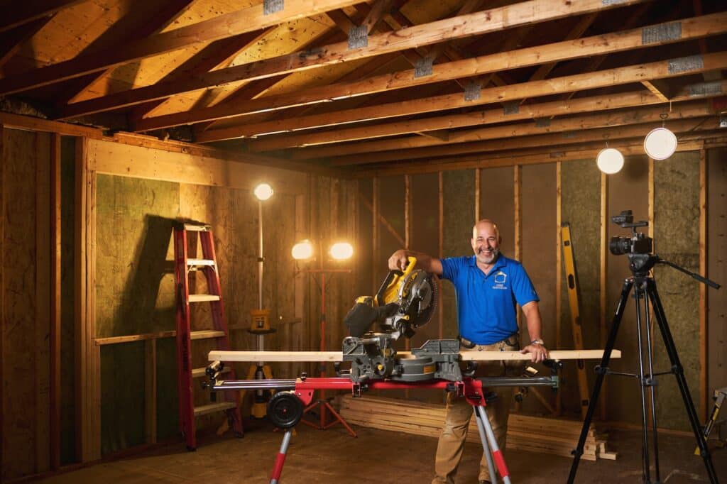 ©JessicaDeeks_20240709_HomeRenoVisionDIY_JeffThorman_0086-JD - House Work Jeff Thorman from HomeRenoVisionDIY in a workshop, smiling with a miter saw and lumber, surrounded by exposed framing and a camera.