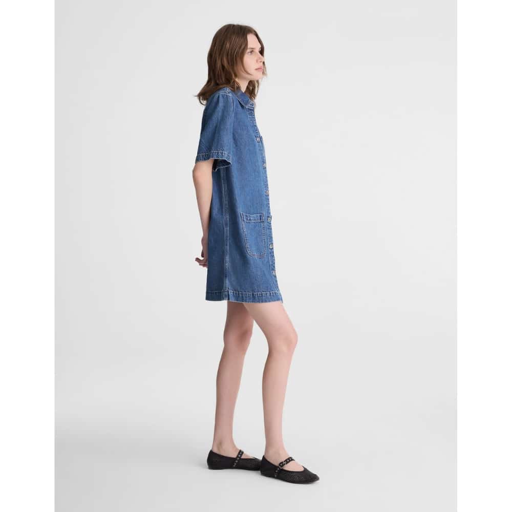 Side view of a woman wearing a knee-length blue denim shirt dress with short sleeves, front buttons and pockets.
