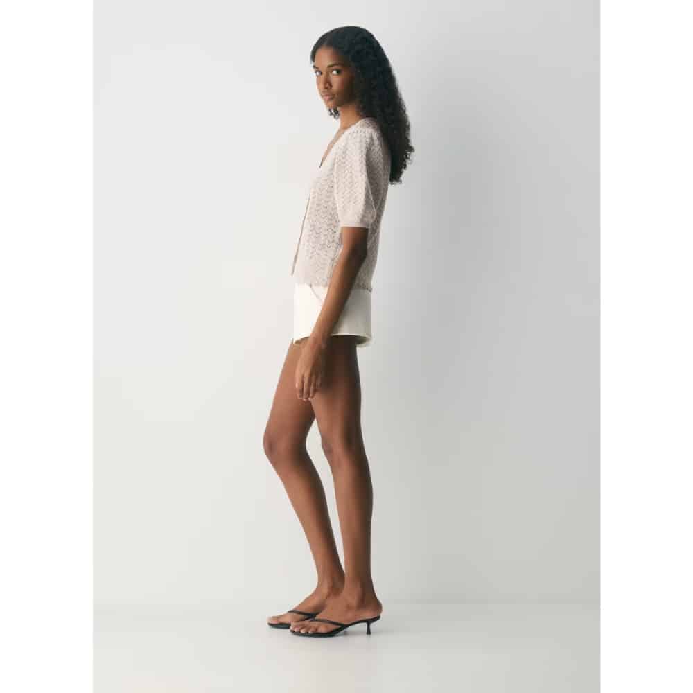 Side view of a woman in a white lace blouse, white shorts, and black flip-flops, standing against a light gray backdrop.