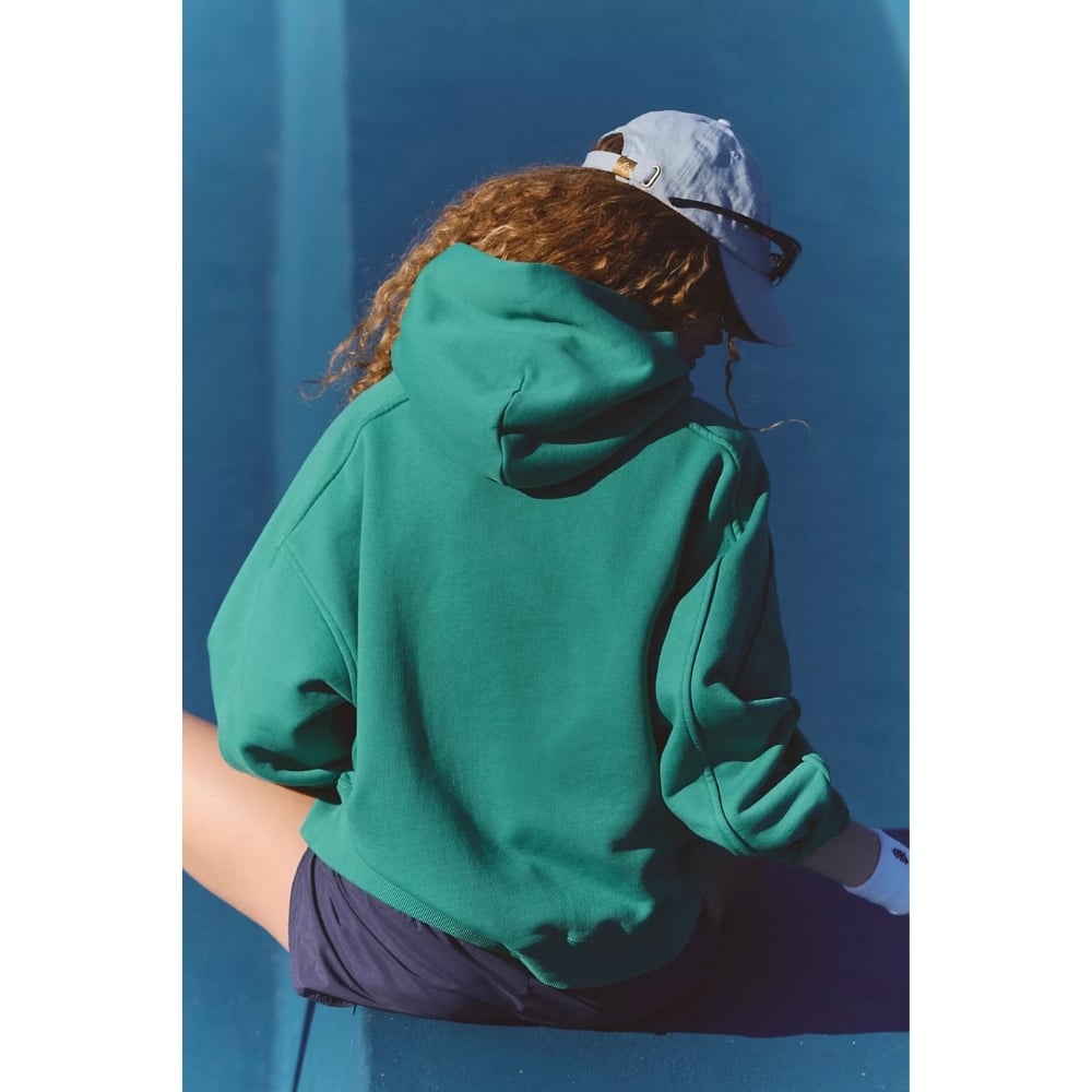Back view of a person in a green hoodie and light blue cap, seen against a blue wall outwardly.