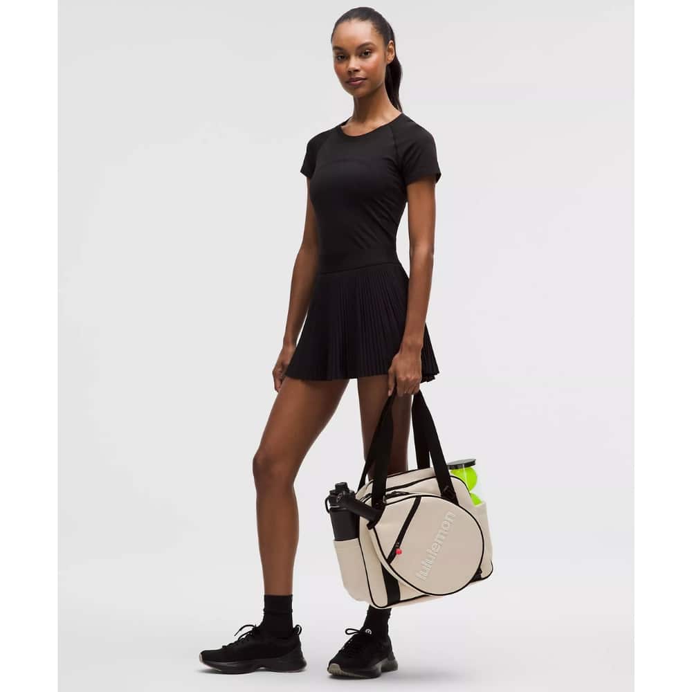 Model wearing a black fitted tee and pleated black mini skirt, carrying a beige gym bag with black straps and a neon bottle inside.
