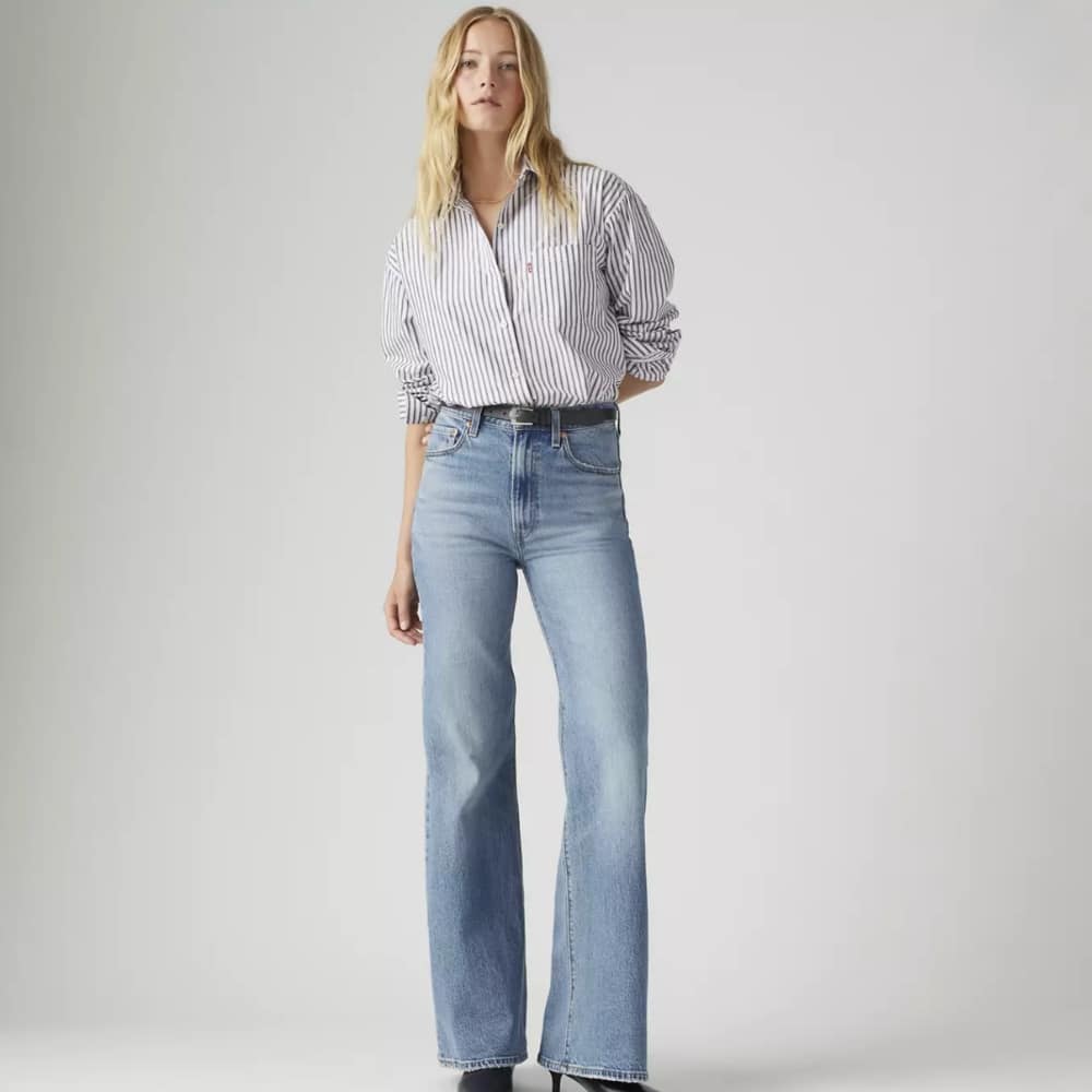 Female model wearing a striped button-down shirt tucked into high-waisted light-blue wide-leg jeans. (informative)