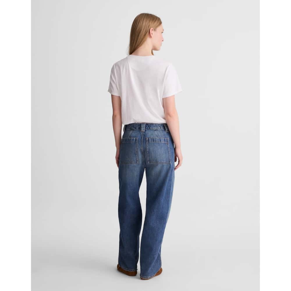 Back view of a person wearing a white t-shirt and high-waisted blue wide-leg jeans, standing against a light gray studio background, brown shoes visible.