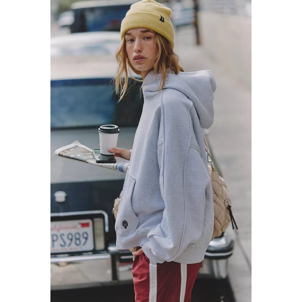 Young woman wearing a gray hoodie and yellow beanie walks on a city street, holding a takeout coffee and a newspaper on a tray, with a parked car behind her and a beige backpack over her shoulder.