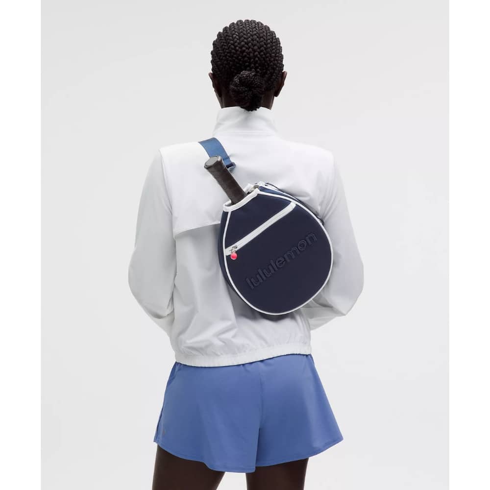 Back view of a person in a white jacket and blue shorts wearing a round navy sling bag with a blue strap and white zipper, seen from behind