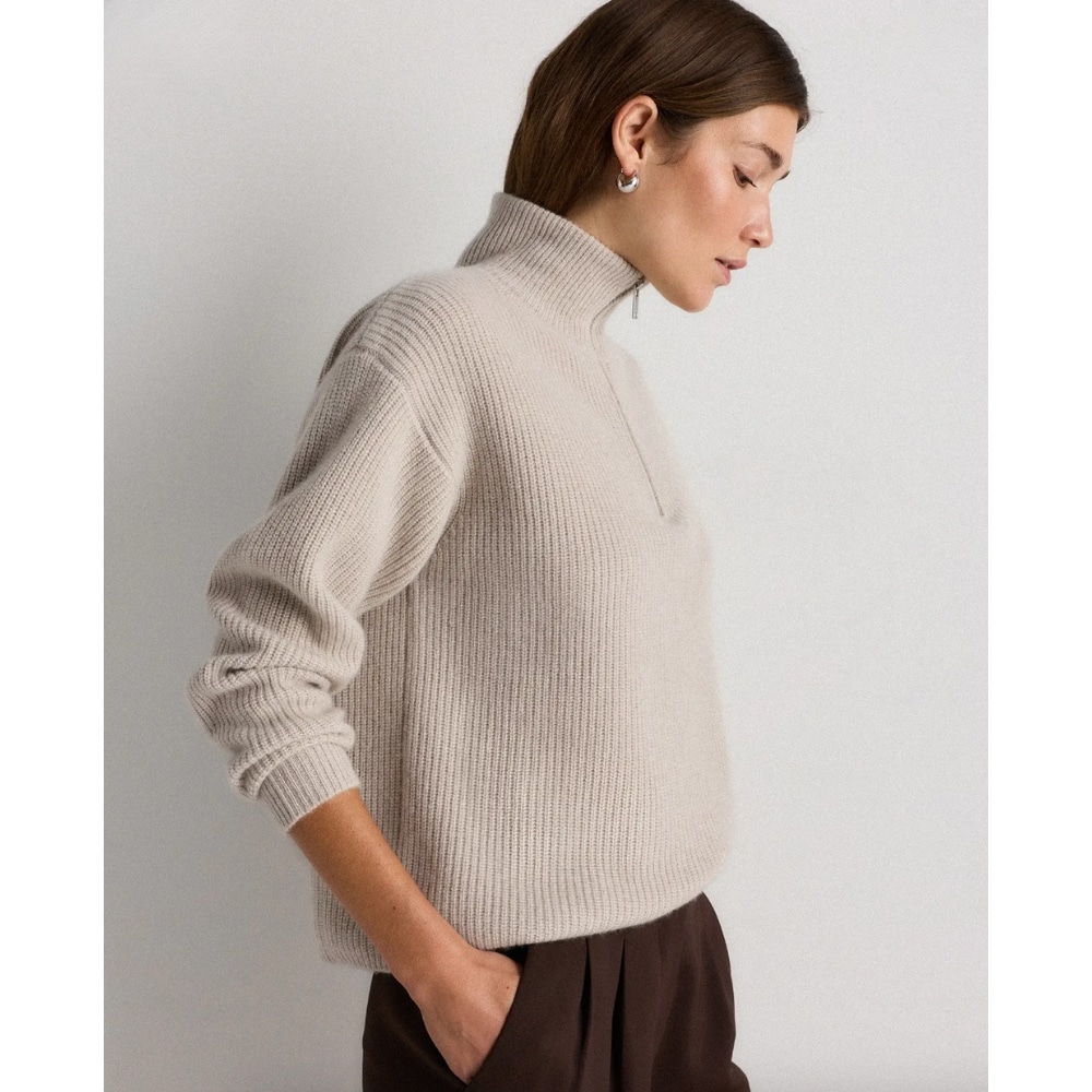 Woman in a beige chunky knit turtleneck sweater, side profile, hands in brown trousers pockets.
