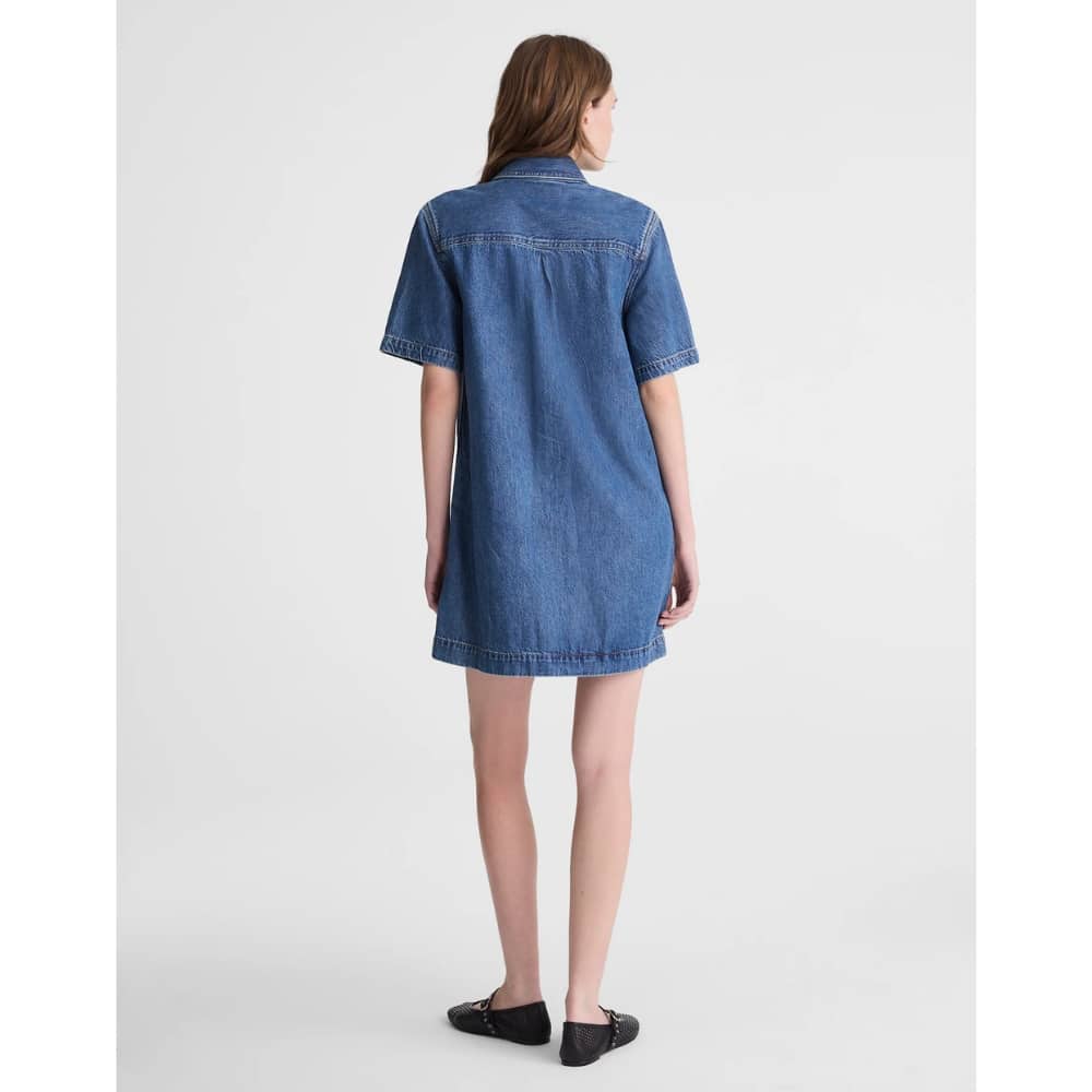 Back view of a person wearing a knee-length blue denim shirt dress with short sleeves and black flats, standing against a light gray background.