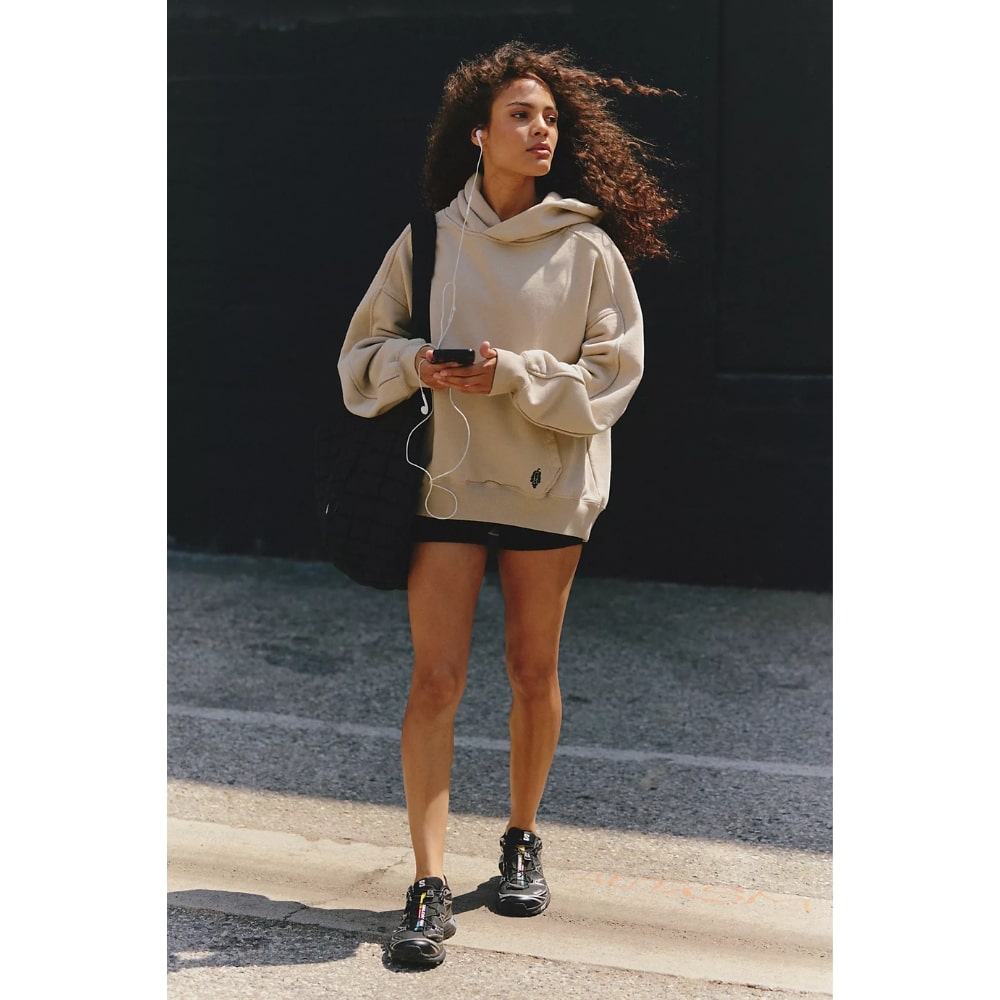 Woman walking outdoors in a beige hoodie, black shorts, and sneakers, listening to headphones while holding a phone and carrying a black bag.