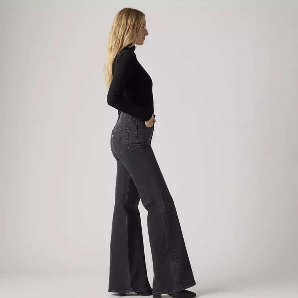 Side view of a woman wearing a black turtleneck and dark gray flared jeans, hands in pockets.