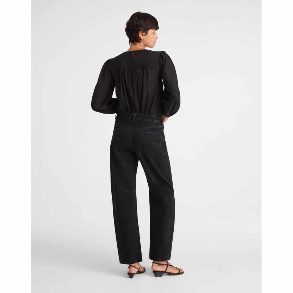 Person viewed from behind wearing a black, long-sleeve gathered-back blouse paired with black high-waisted jeans and strappy sandals.