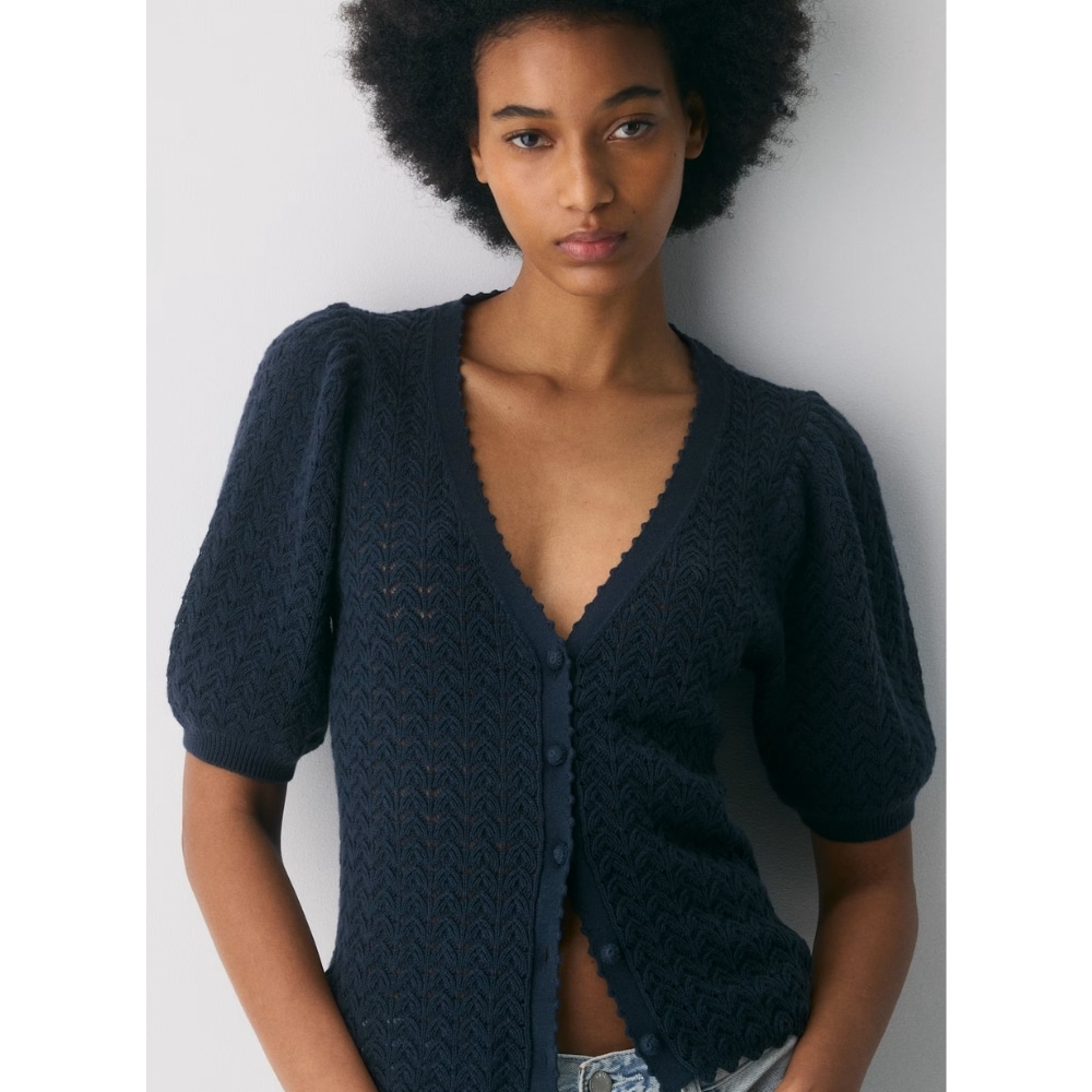 Model wearing a dark navy knitted cardigan with puff sleeves and a deep V-neck, standing against a light wall.