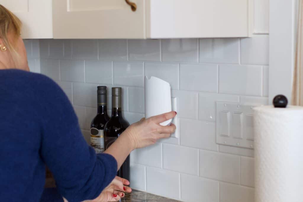 Photo 2026-02-12 1 05 41 PM - House Work Sarah plugging a fruit fly trap into a wall on a white subway tile kitchen backsplash, next to wine bottles and a light switch.