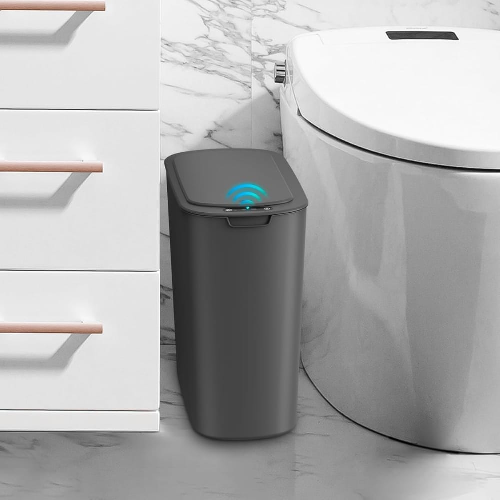 Gray smart trash can with a blue wireless signal icon on the lid, placed in a bathroom beside a toilet and white vanity drawers.
