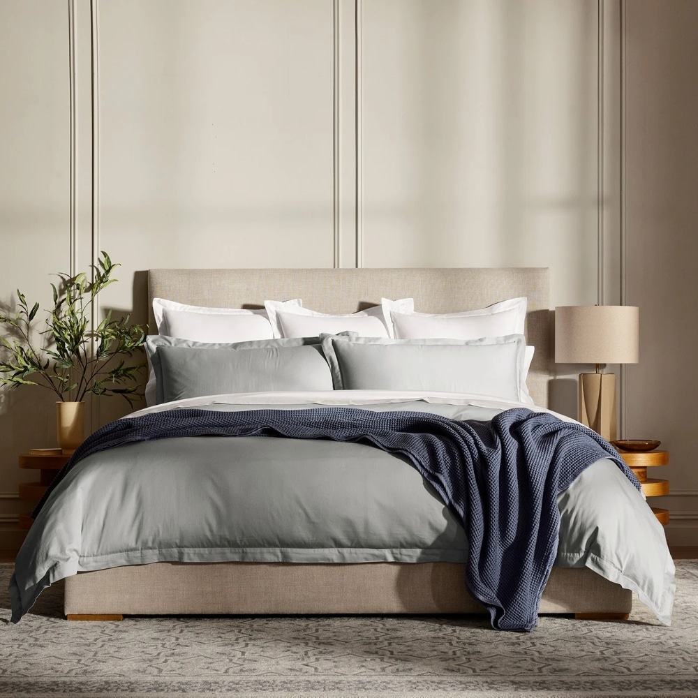 A neatly made bed with a beige upholstered headboard, white and gray linens, and a navy knit throw draped across the foot of the bed, in a calm bedroom.