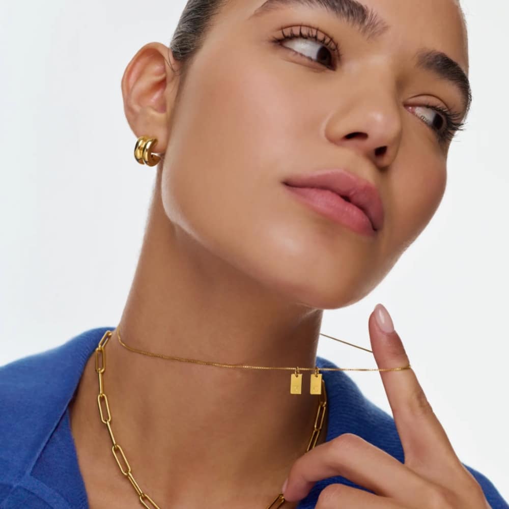 Close-up of a model wearing gold jewelry: a thin choker with small rectangular pendants and a chunky chain necklace, paired with hoop earrings and a blue garment.