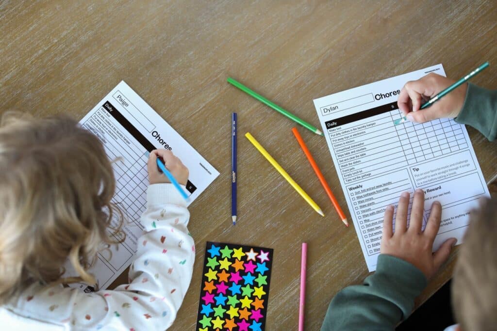2178322_HWChoreChart11-14_Photos_7_030124 - House Work Two young kids sitting at a table filling out a summer chore chart printable with colored pencils and star stickers.