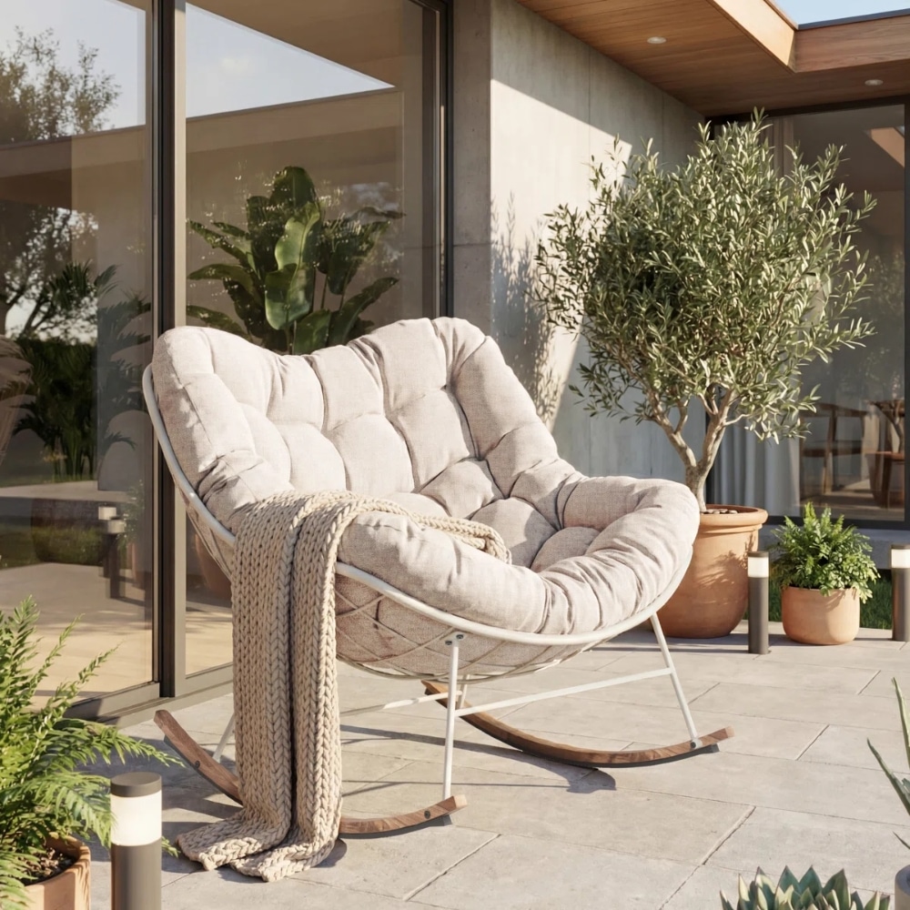 Cream tufted rocking chair on a sunny patio, with a chunky knit blanket draped over it and potted plants nearby by a glass-walled house.