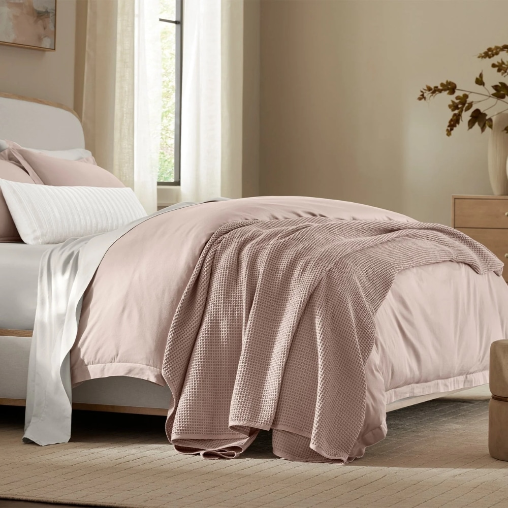 Bed made with blush pink comforter and textured pink throw blanket in a bright bedroom point of view.