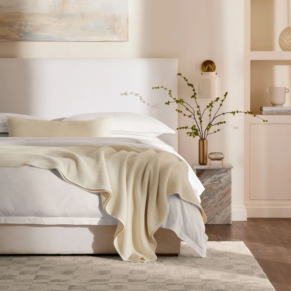 Creamy neutral-toned bedroom with a white bed, beige knit throw, and a marble nightstand topped with a brass vase of greenery.