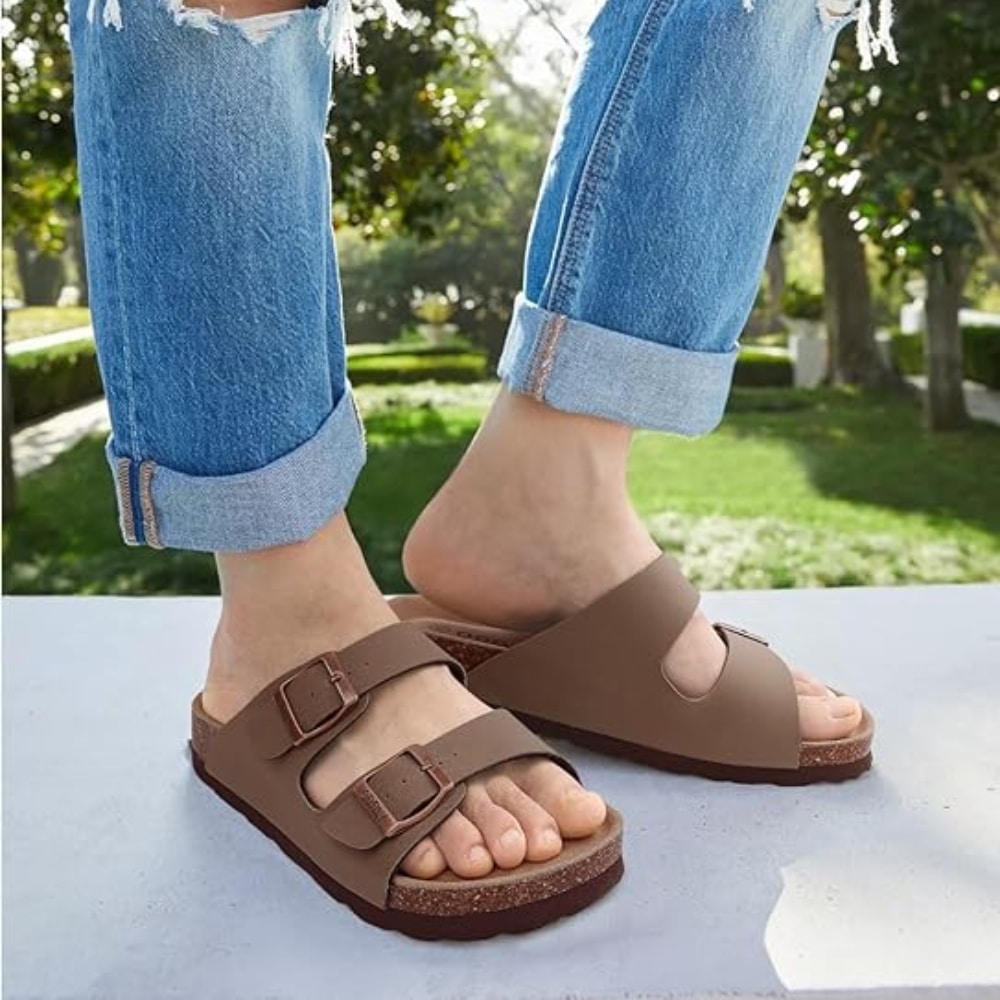 Person wearing brown double-buckle sandals with rolled denim jeans, standing outdoors on a light surface.