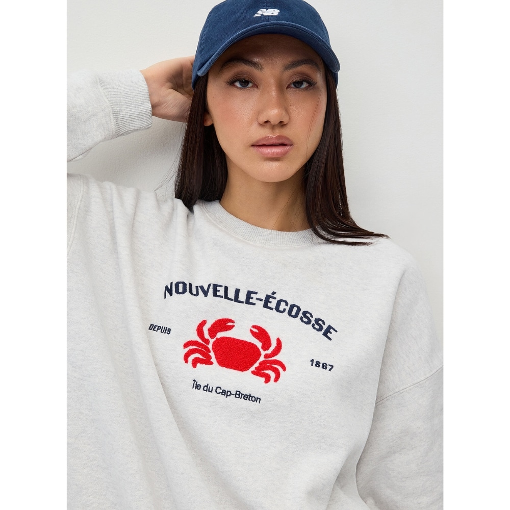 Portrait of a woman wearing a light gray sweatshirt with a red crab graphic and the text 'NOUVELLE-ÉCOSSE', plus a blue New Balance cap.