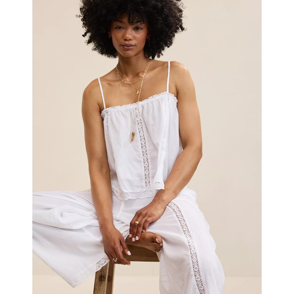 Woman with natural curly hair sits on a wooden stool, wearing a white strap top and white pants with lace trim, posing against a beige background.