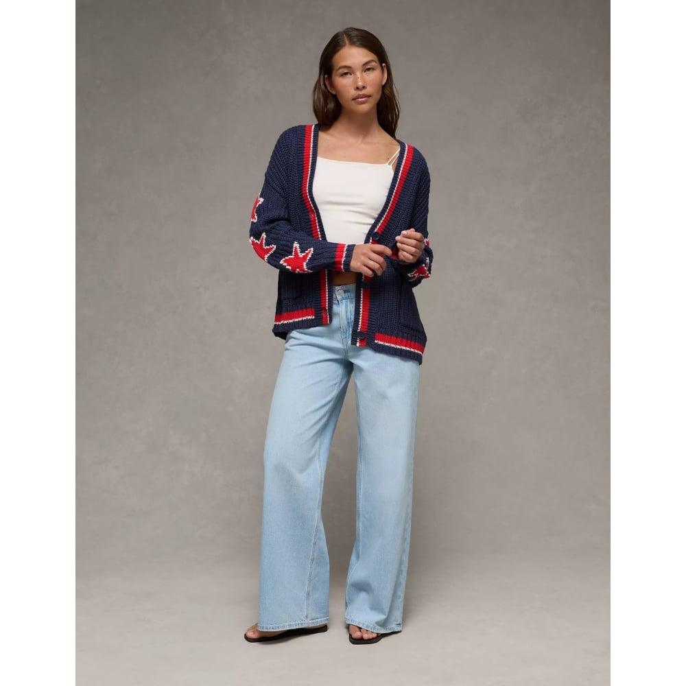 Woman wearing a navy knitted cardigan with red star patterns over a white tank top and light blue wide-leg jeans, standing against a gray studio backdrop.