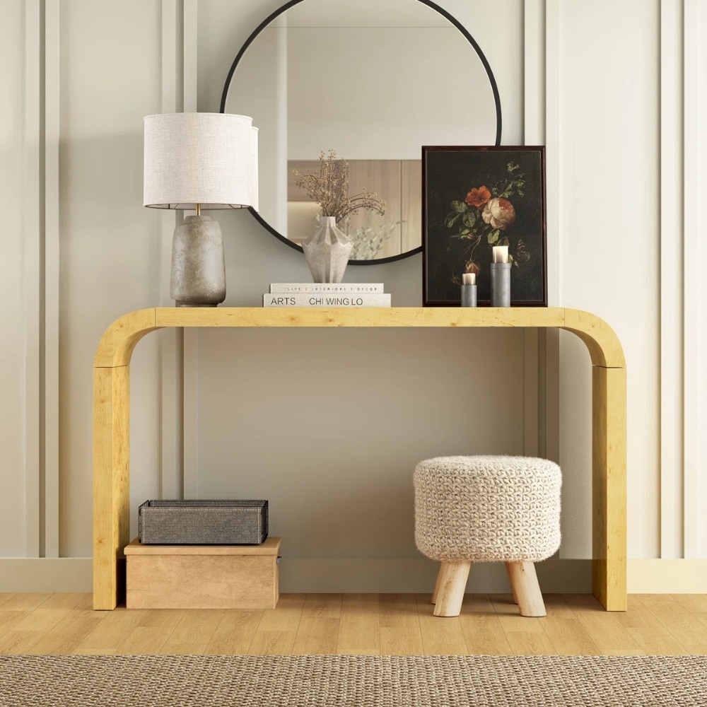 Wood console with rounded legs under a large round mirror, featuring a lamp, vase, books, framed floral art, candles, and a knit stool.