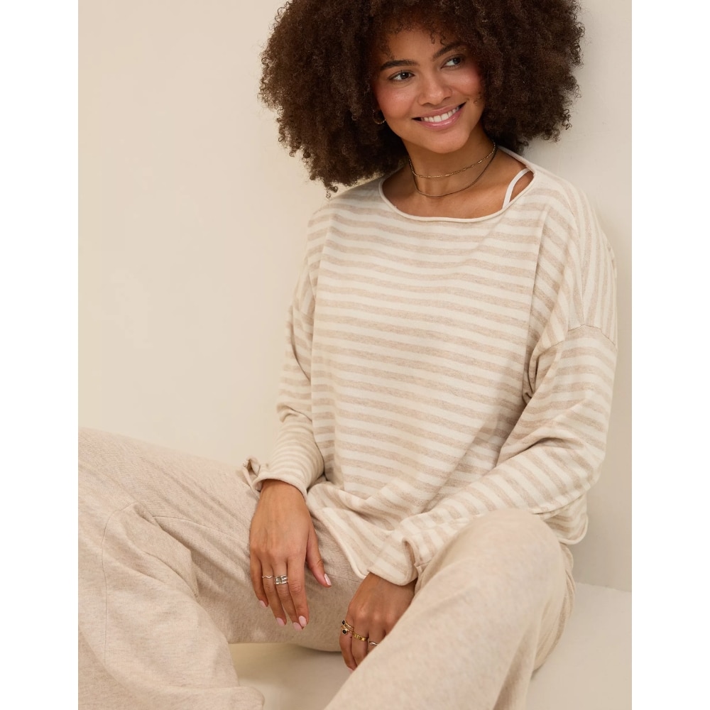 Woman with curly hair wearing a beige striped sweater and light beige pants, sitting against a neutral background and smiling.
