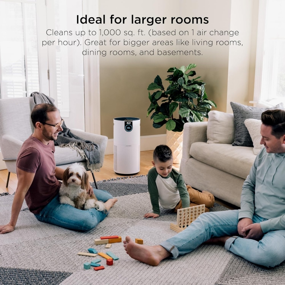 White cylindrical air purifier in a bright living room as a family sits on the rug with a dog and toys nearby.