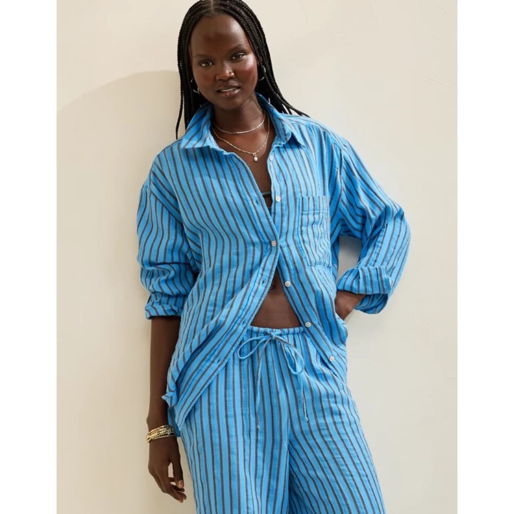 Woman in a blue striped matching shirt and pants set, shirt open to reveal midriff, posing casually against a light wall.