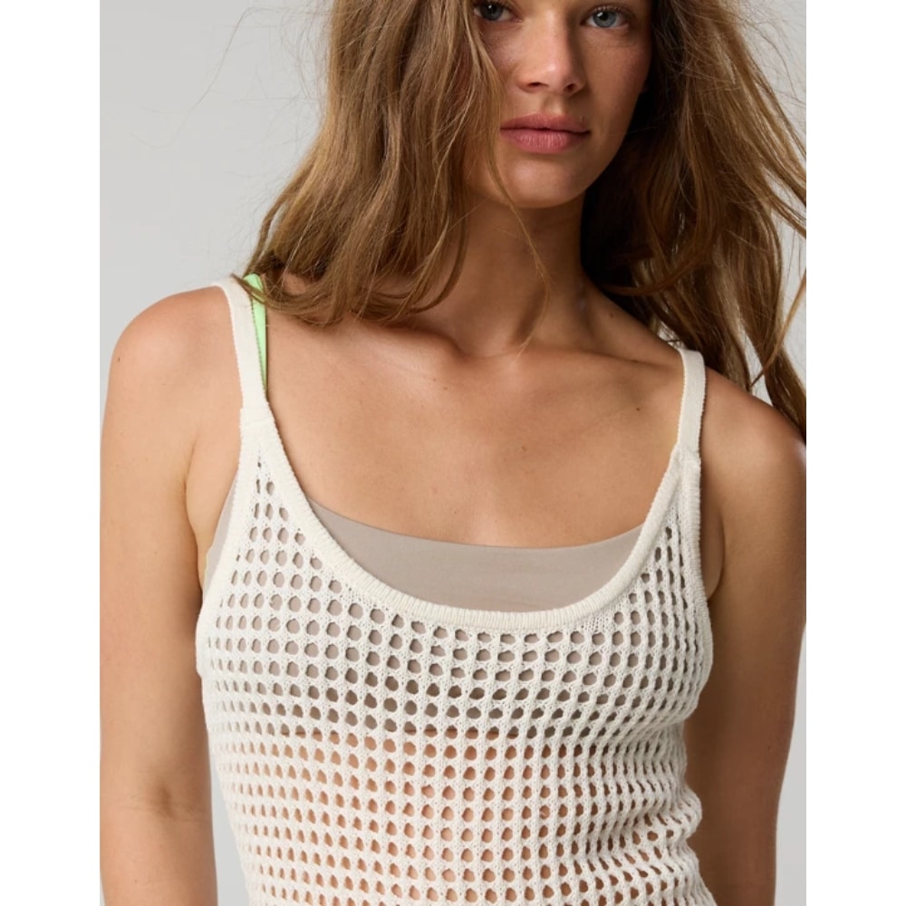 Close-up of a woman wearing a white crochet mesh tank top over a beige bra, with light brown hair partially covering her face.