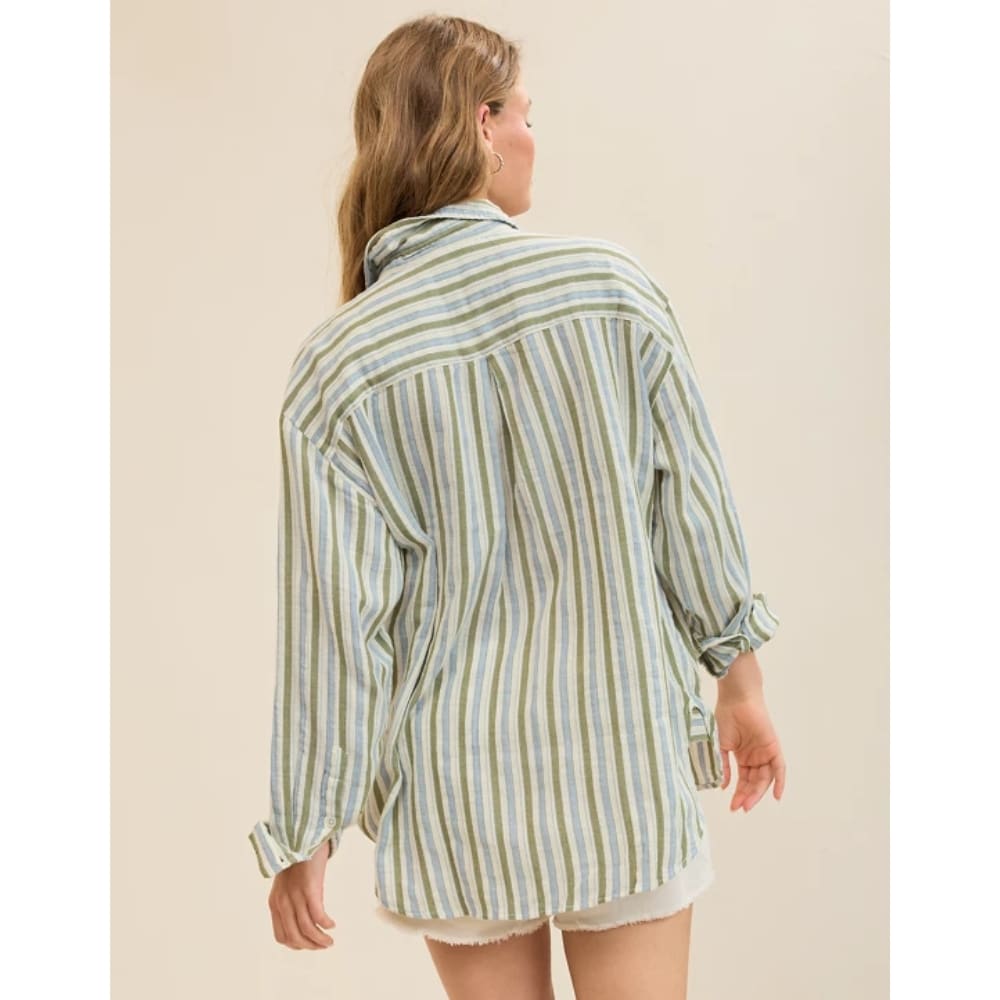 Back view of a person wearing a loose striped shirt in pastel greens and blues, with rolled cuffs, over white shorts, against a beige background.