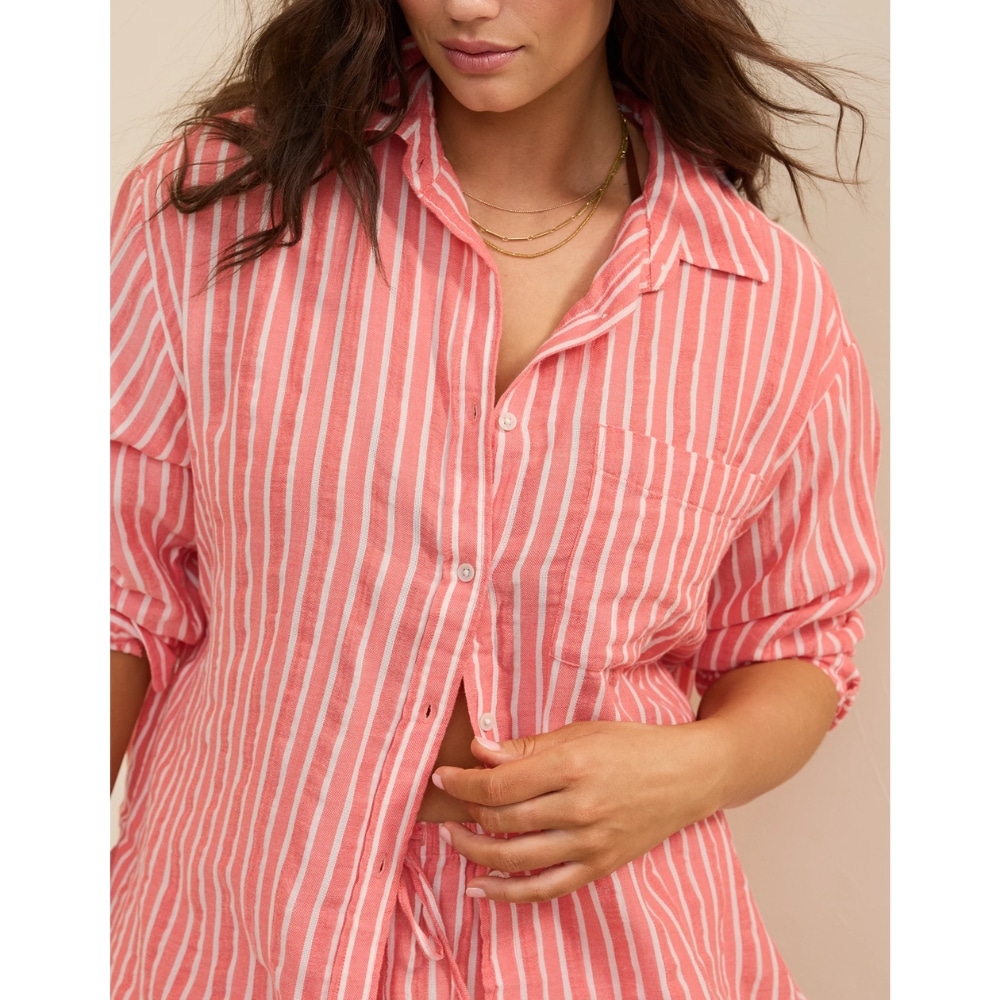 Woman wearing a pink and white striped button-up shirt, unbuttoned at the waist, adjusting the hem with her hand; gold layered necklace visible.
