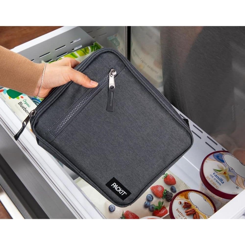 Gray PackIt insulated lunch bag being placed into a freezer drawer with frozen items visible in the background.