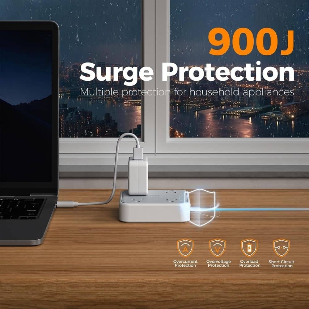 White surge protector with a plug connected to a laptop, labeling reads '900J Surge Protection' with protection icons below; device sits on a wooden desk by a window.