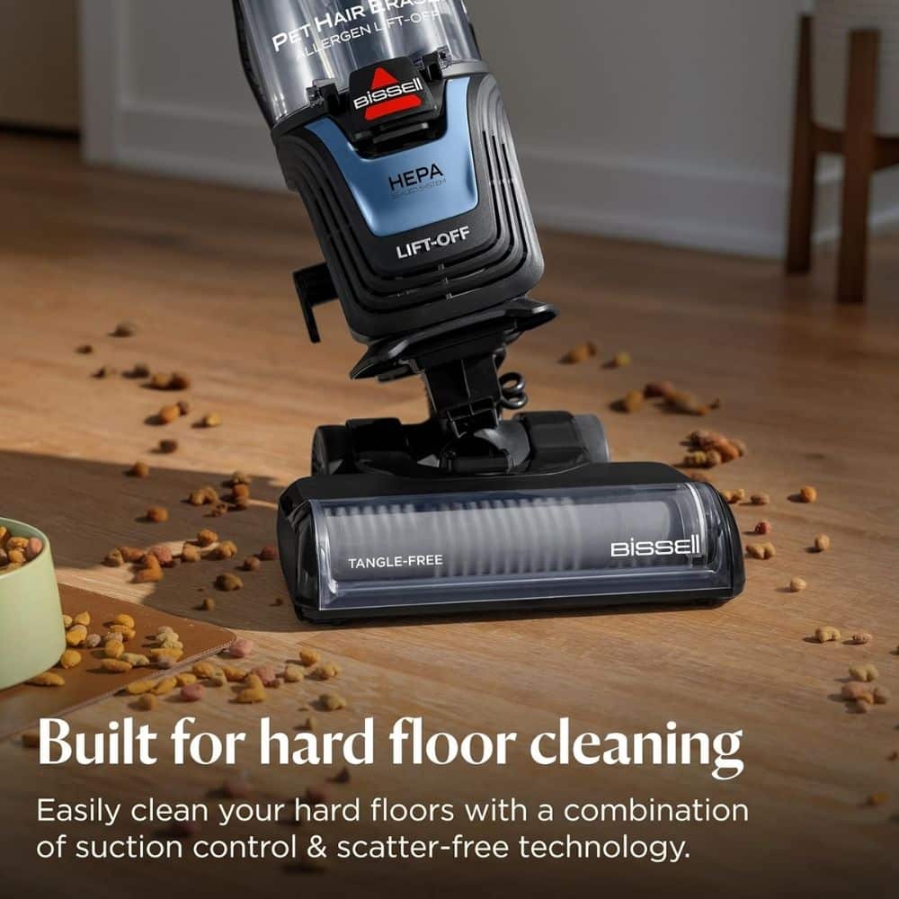 Bissell upright vacuum with a tangle-free brush head on a hardwood floor, sweeping scattered pet kibble on the floor.