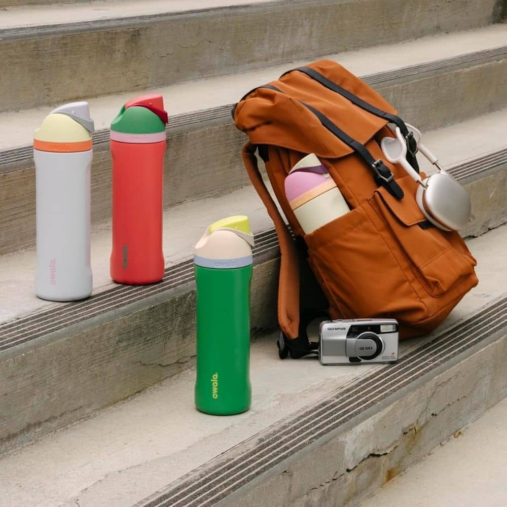 Colorful insulated bottles lined up on concrete steps next to an orange backpack and a small silver Olympus camera with headphones attached to the bag