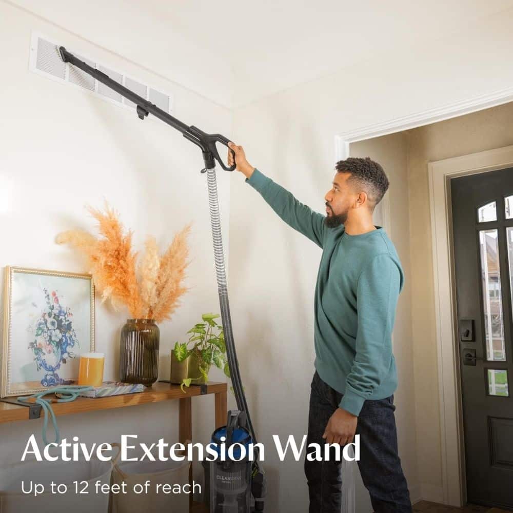 Man uses a long-extension wand to clean a ceiling vent in a living room, reaching high into the vent area with a vacuum hose attached.