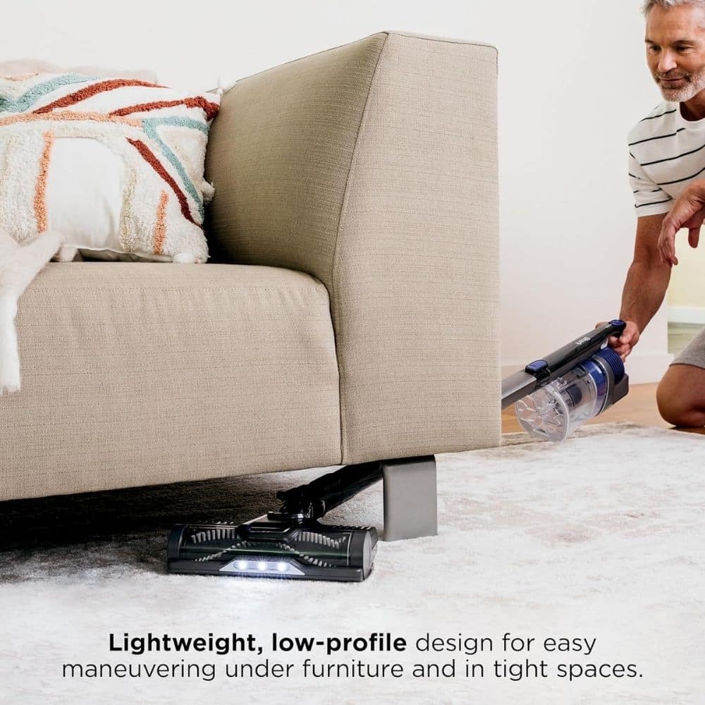 Person vacuuming under a beige sofa with a low-profile, lightweight vacuum designed for maneuvering in tight spaces.