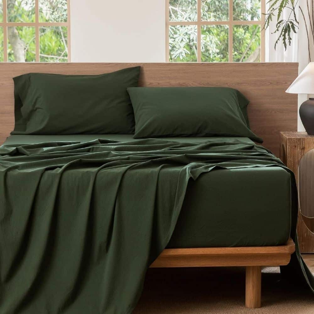 Bed made with dark green sheets and two matching pillows against a wooden headboard, with a nightstand and lamp to the right.