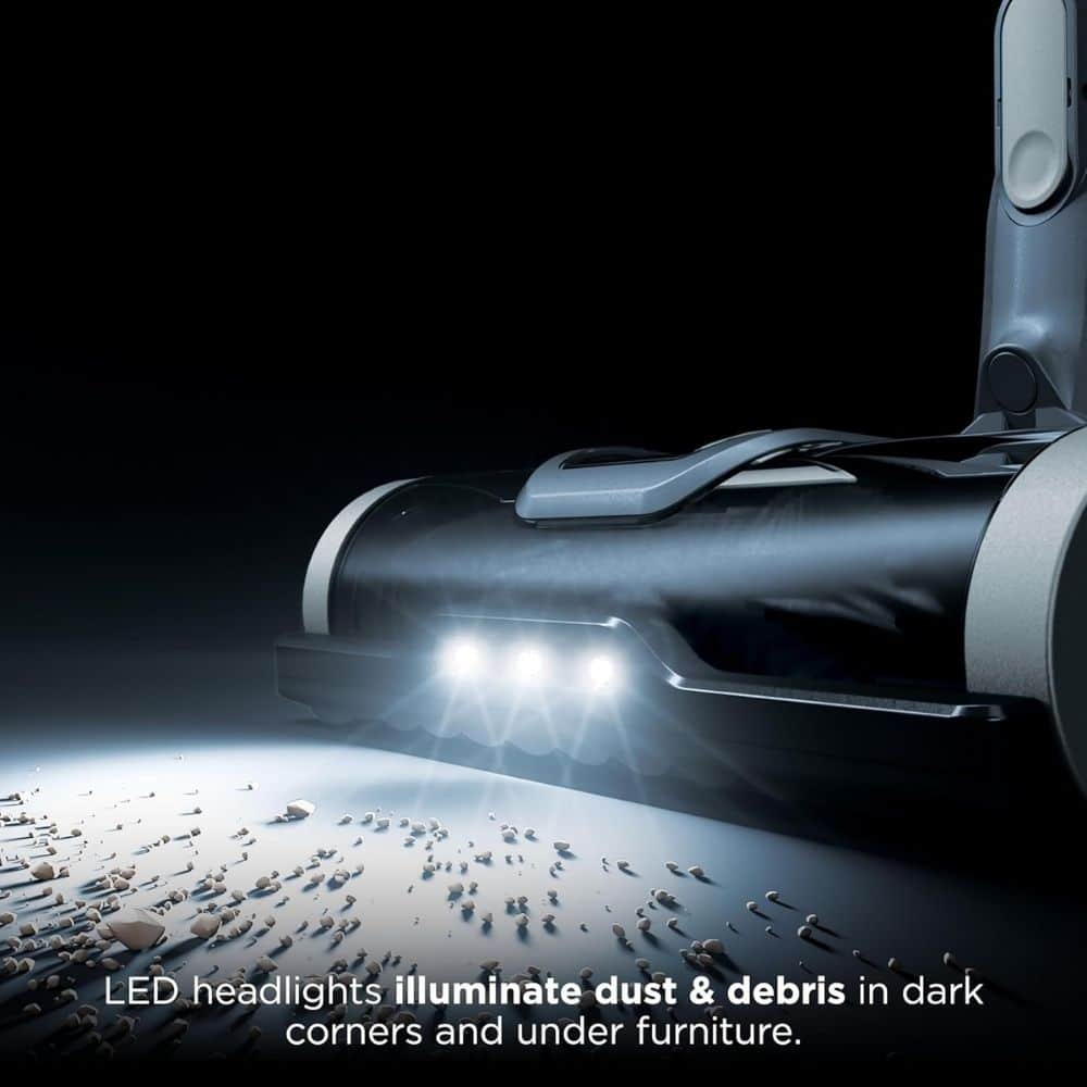 Cordless vacuum head with LED lights illuminating dust and debris on a dark floor near furniture edge.