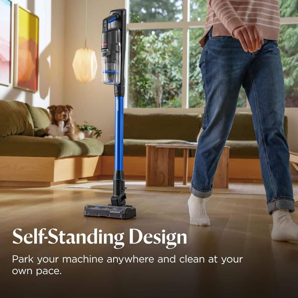 Upright vacuum cleaner stands on a living room floor with a person nearby and a dog on the sofa; overlay text reads 'Self-Standing Design'.