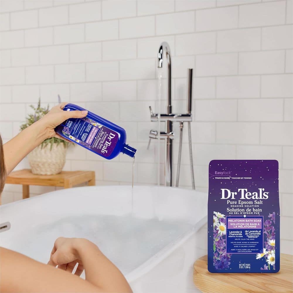 Person pouring liquid from a blue bottle into a white bathtub beside a blue Dr Teal's bath soak pouch on a wooden tray/stand
