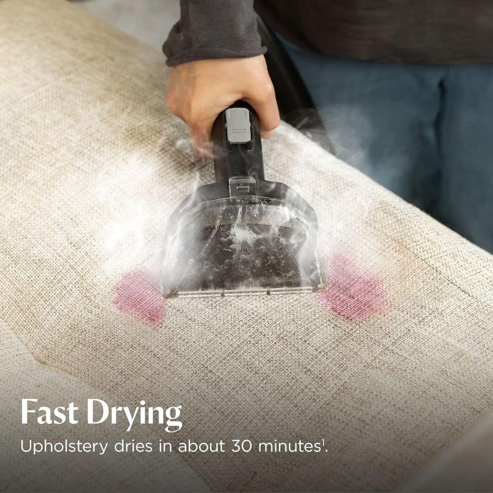 Person vacuuming a beige sofa with a steam/cleaning tool, revealing a pink stain and mist from the device. Text on image: "Fast Drying" and "Upholstery dries in about 30 minutes".