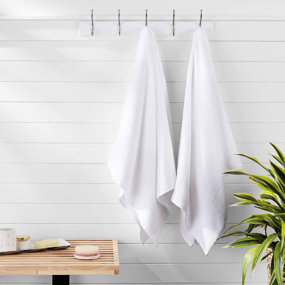 Two white towels hang from a metal rack on a white paneled bathroom wall, with a wooden bench of toiletries nearby and a green plant to the side.